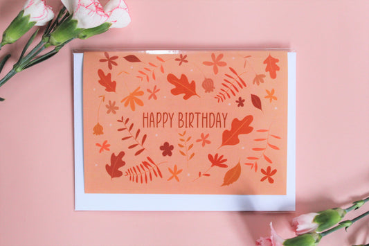 "Happy Birthday" greetings card with peach background with an autumnal colour foliage pattern