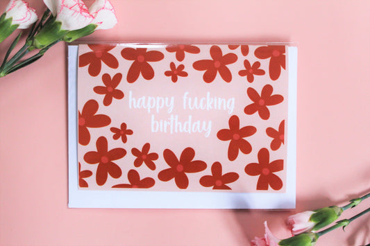"Happy f***ing birthday" greetings card on pink background with terracotta coloured flowers.